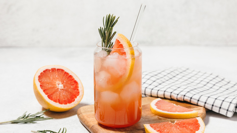 Sparkling Grapefruit soda in a tall glass with ice, rosemary, and glass straw surrounded by grapefruit slices