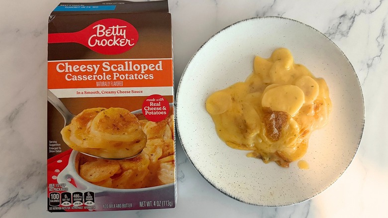 Box of Betty Crocker Cheesy Scalloped Potatoes next to scalloped potatoes on white plate