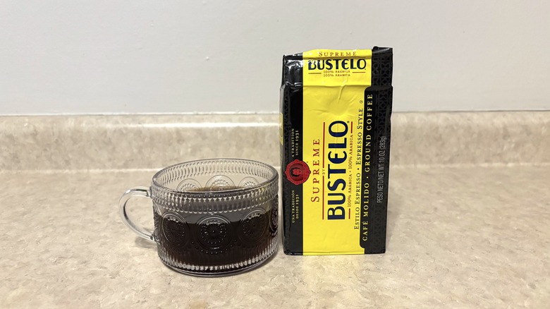 Cafe Bustelo supreme ground coffee in brick and glass mug on neutral background.