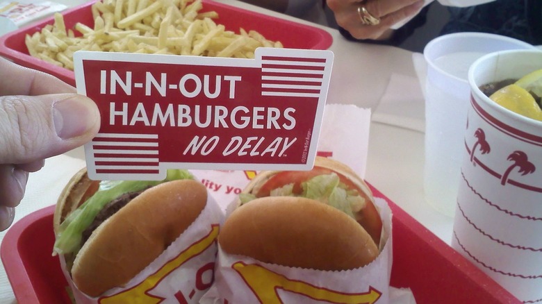 Fingers holding an In-N-Out Hamburger No Delay bumper sticker over a tray with two paper-wrapped burgers, soft drink, and a basket of fries