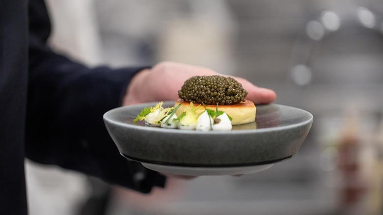A server's hand holding a plate of delicately arranged pancake, dollops of sauce, and a giant helping of caviar