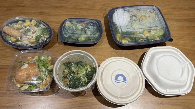Seven salads in closed containers on a wooden surface.