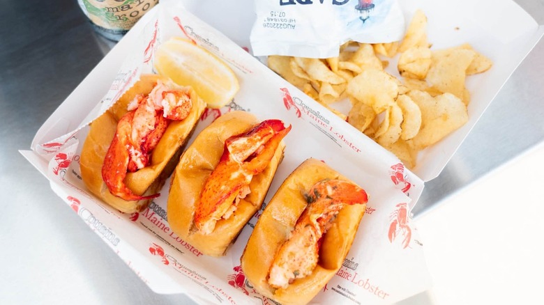 Three lobster rolls from Cousins Maine Lobster sitting in a basket next to Cape Cod potato chips and a root beer.