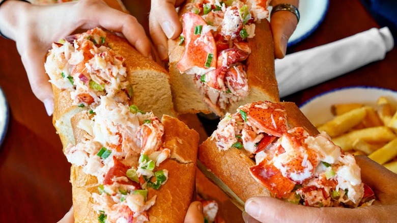 A group of hands holding Legal Sea Foods lobster rolls up to each other brimming with lobster meat and green onion.