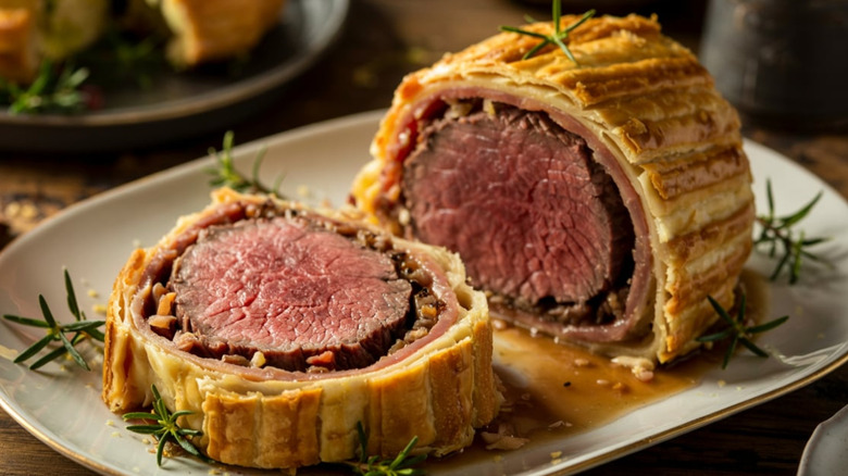 Beef Wellington with a puff pastry crust, sliced to show pink beef tenderloin inside, on white plate garnished with rosemary