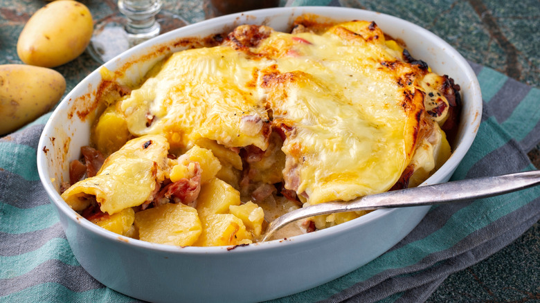 Oven-baked Tartiflette in casserole dish on table cloth next to loose potatoes