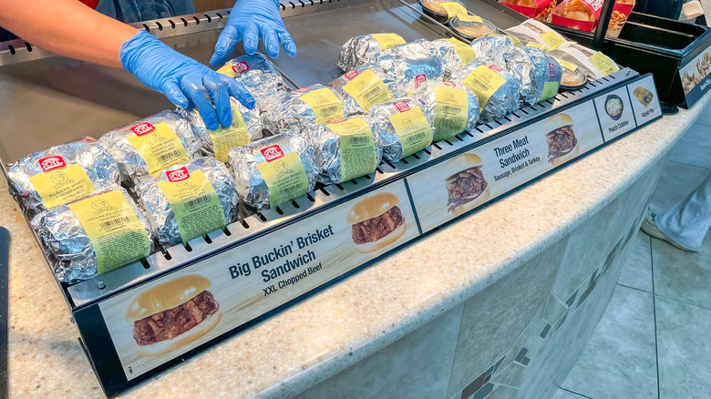 Rows of foil-wrapped, yellow-labeled sandwiches are placed in a display by a pair of gloved hands. A sign at the front of the display advertises the "Big Buckin' Brisket Sandwich" and "Three Meat Sandwich."