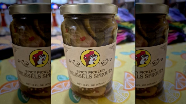 A glass jar with a beige label reading "Spicy pickled brussels sprouts" with Buc-ee's logo sits on a table with a yellow tablecloth depicting various multicolored sliced citrus fruits.