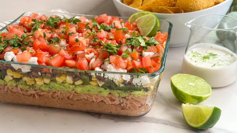 Elevated 7-Layer Dip in clear dish with limes and a creamy white sauce on the side