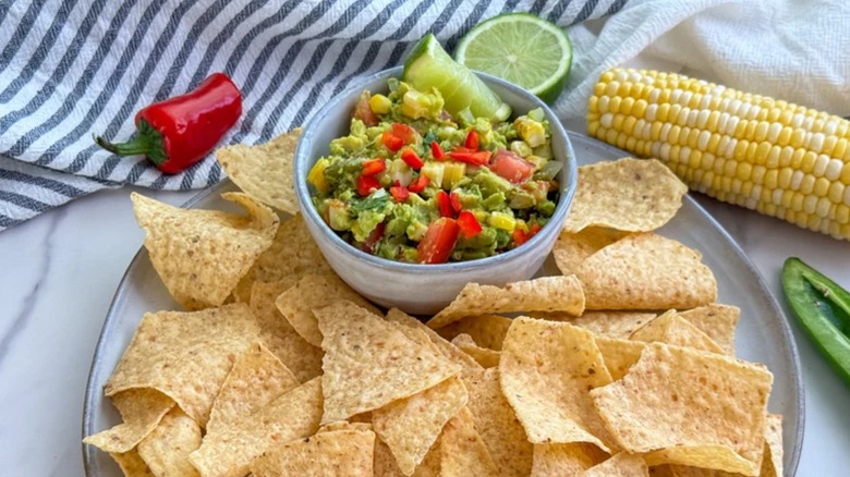 Corn and avocado guacamole served in a bowl topped with chopped red peppers on a plate with chips and corn cob and a red pepper on the side