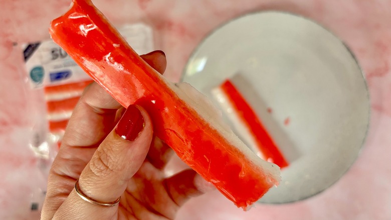Hand holding imitation crab leg above plate and bag of imitation crab on pink background
