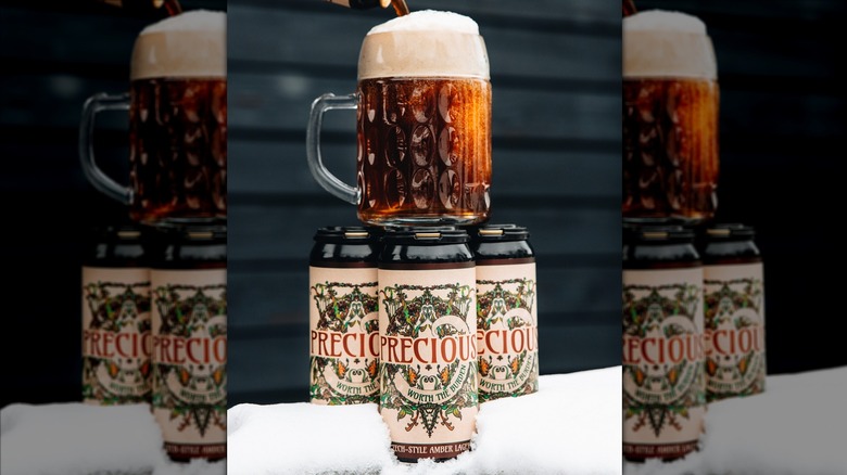 Three cans of Burial Precious Czech-style Amber Lager on snow with a mug of beer on top of them