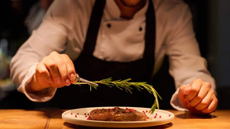 The hand of a chef putting aromatic herb on top of roasted meat.