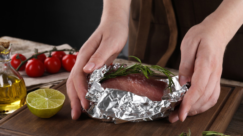 hands holding foil packet of raw meat on wood cutting board with other ingredients in background