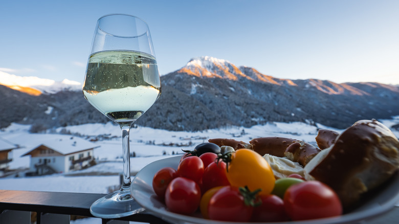 Hotel balcony table shows white wine, tomatoes, olives, cheese, and bread in front of snowy mountains