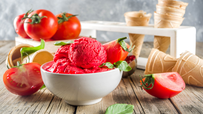 Tomato ice cream in a bowl with fresh tomatoes and basil and leaves and cones behind