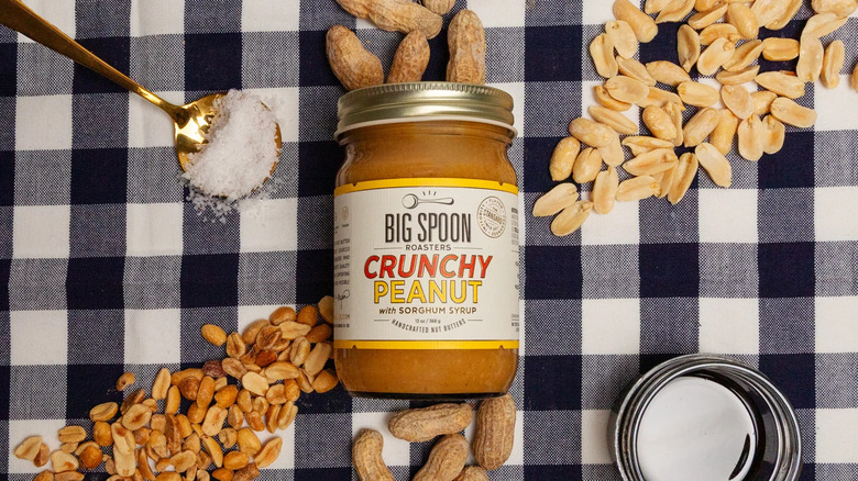 Overhead view of Crunchy Peanut Butter jar on tablecloth, surrounded by two types of peanuts and spoonful of salt