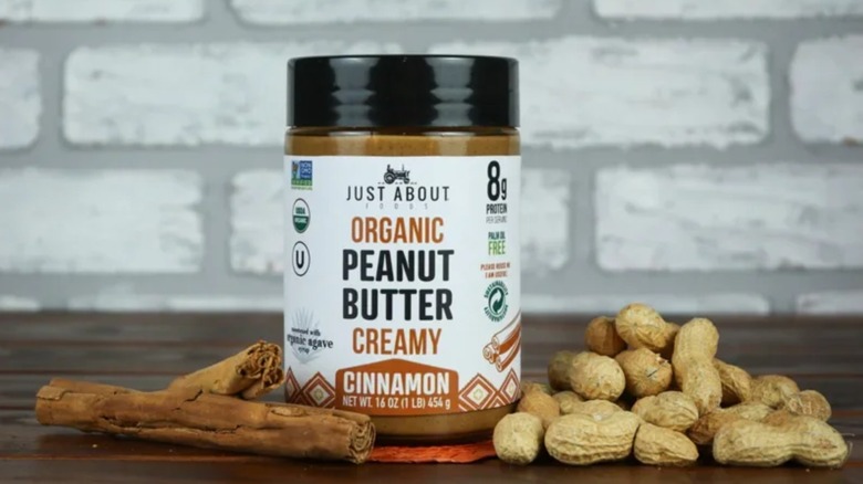 Jar of Just About Foods cinnamon peanut butter surrounded by cinnamon sticks and peanuts