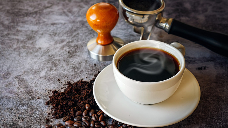 White mug with black coffee on white saucer with whole coffee beans and ground coffee nearby.