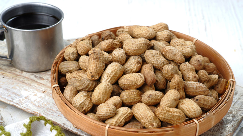 A whicker boal of peanuts in the shell, next to a tin cup, on a table