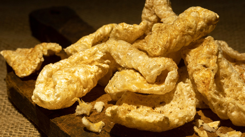 A close-up image of a pile of pork rinds on a wooden cutting board against a stark background.