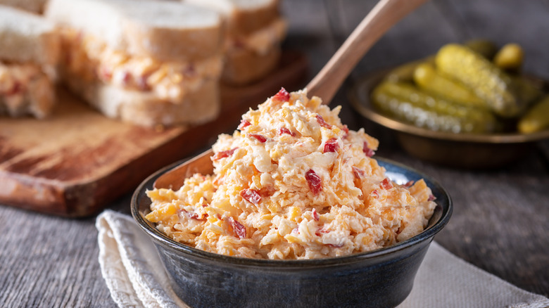 A bowl of home-made pimento cheese in front of a cutting board with pimento cheese sandwiches next to a plate of cornichon pickles