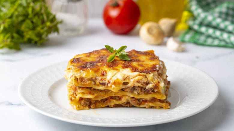 Piece of lasagna on plate in front of garlic, tomato, and herbs