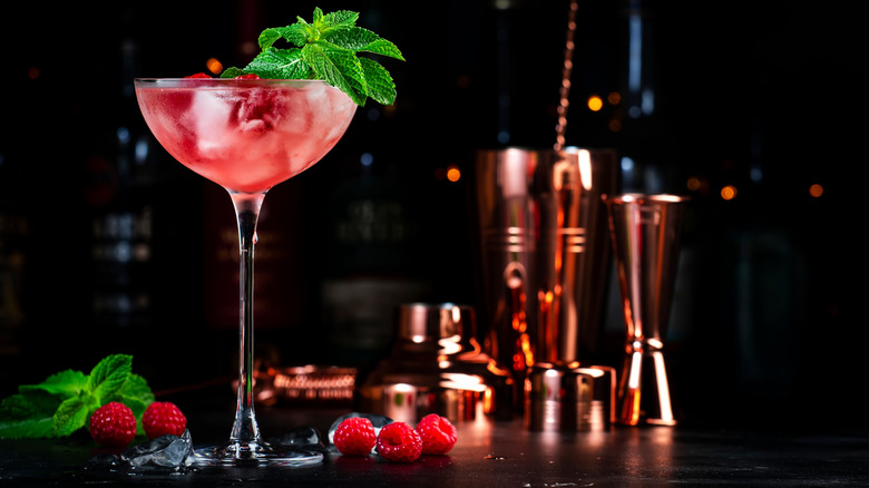 tall pink cocktail with ice and mint with raspberries and cocktail shaker on the side