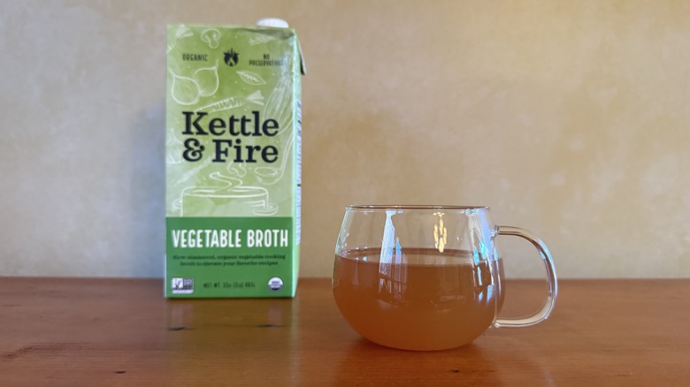 Carton of Kettle & Fire vegetable broth on wooden table with cup of broth