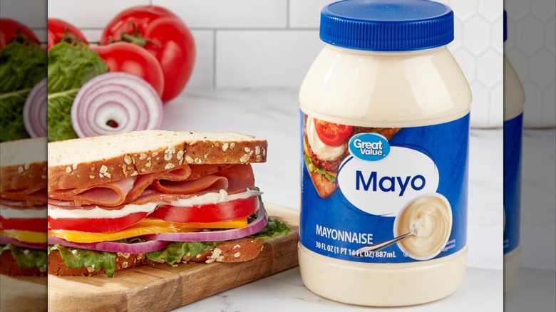 Tub of Great Value Mayonnaise sitting next to a sandwich stuffed with meat, cheese, and veggies, alongside a spread of mayonnaise.