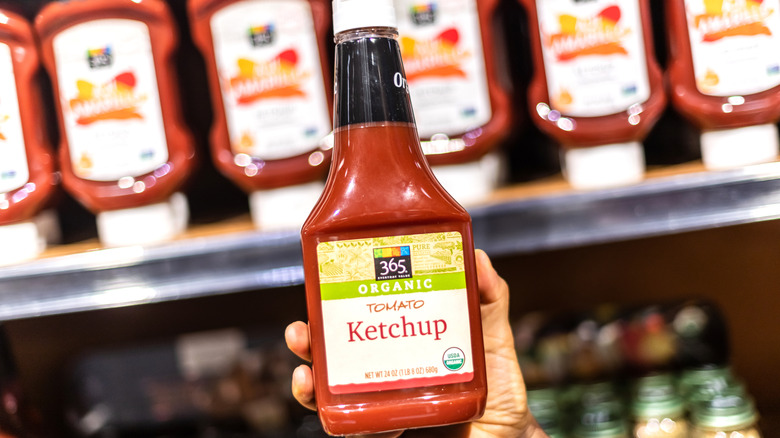 Someone holding up a bottle of 365 Organic Tomato Ketchup with a lineup of ketchup on a shelf behind them.