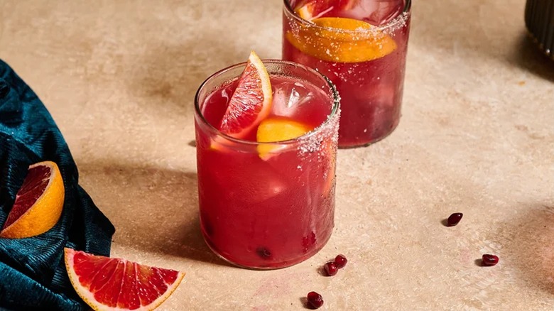 Blood orange and pomegranate margaritas next to blood orange slices and pomegranate seeds on beige surface