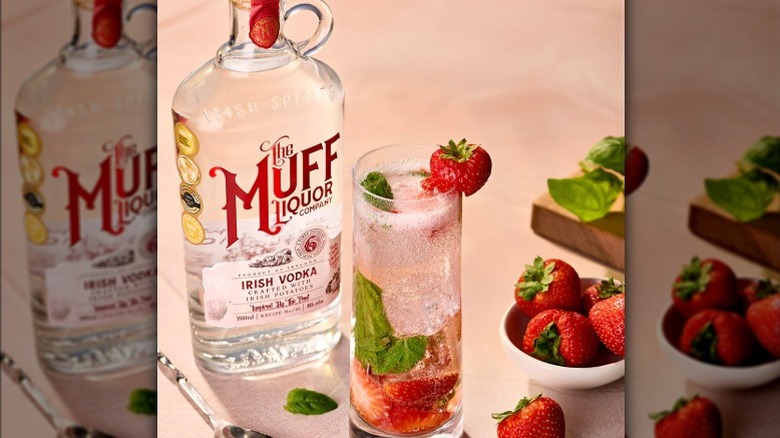 Label view of Muff Irish Vodka bottle next to highball with strawberry garnish, basil and fruit strewn about