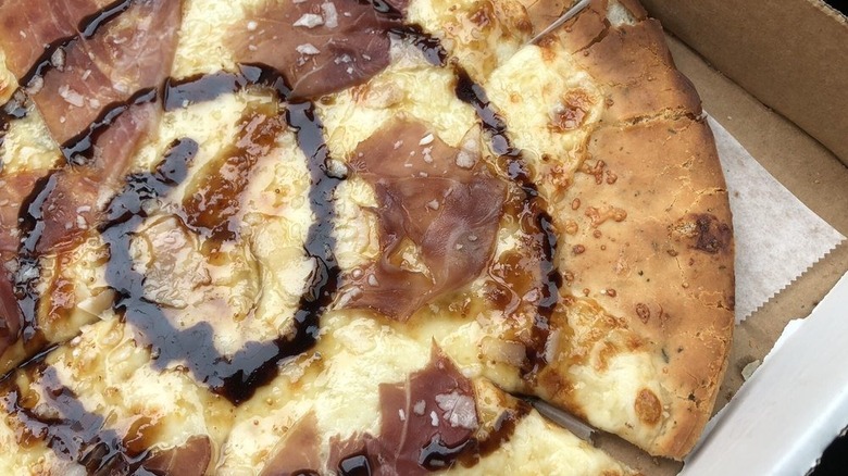 Pizza in box topped with fig preserve, prosciutto, balsamic reduction, and various cheeses