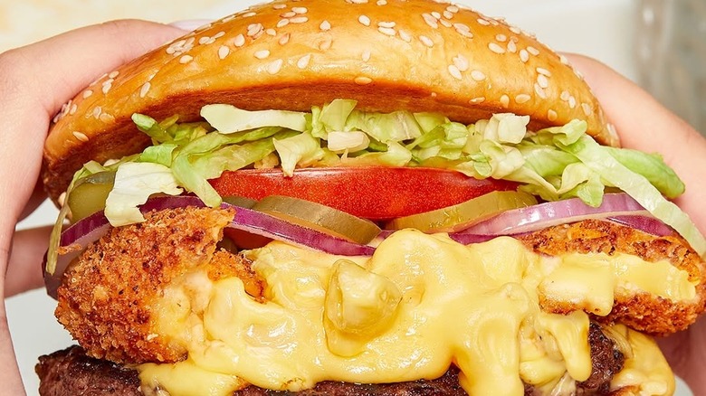 Hands holding a Cheesecake Factory Macaroni & Cheese Burger with red onions, pickles, tomato, shredded lettuce and a fried macaroni & cheese ball.