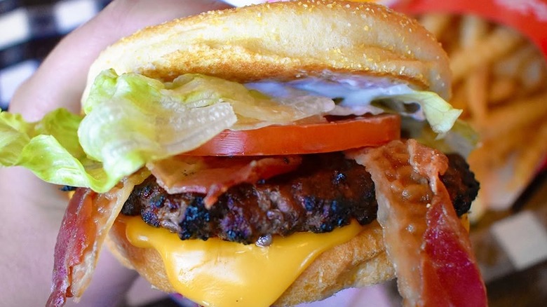 A left hand holding a Portillo's bacon cheeseburger with mayonnaise, lettuce and tomato and fries in the background.