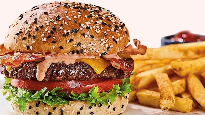 Close-up of Red Lobster's bacon cheeseburger with lettuce, tomato slices and signature sauce on a sesame bun, with seasoned fries and a small ramekin of ketchup on the side.