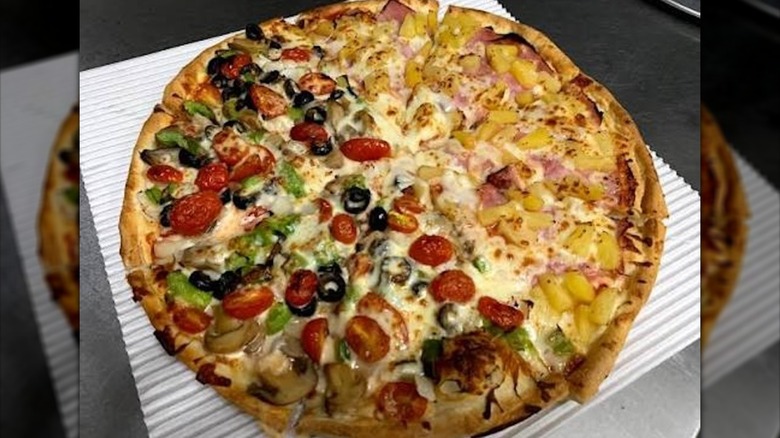 Split pizza with pineapple and ham on one side and pepper, tomatoes, and mushrooms on the other half