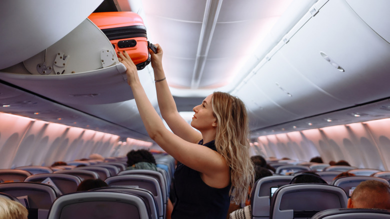 Woman placing hardtop suitcase in overhead compartment on plane