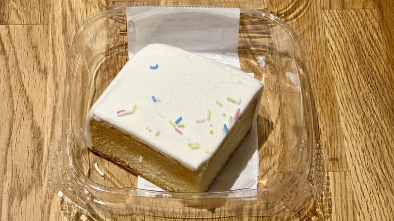 Plastic container of slice of Whole Foods Vanilla Sprinkle Celebration Cake on wooden countertop