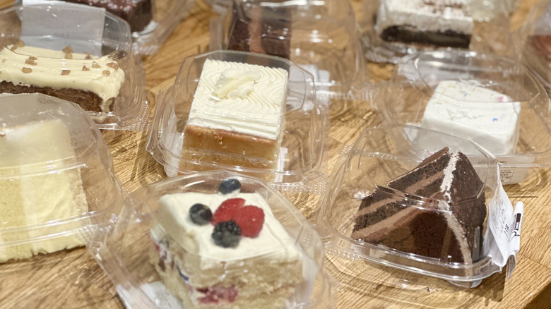 Overhead shot of multiple plastic containers of cake slices from Whole Foods on wooden counter