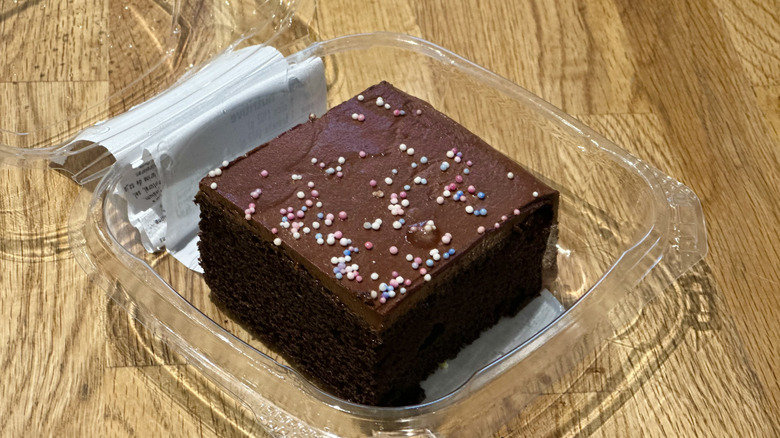 Plastic container of slice of Whole Foods Chocolate Sprinkle Celebration Cake on wooden countertop