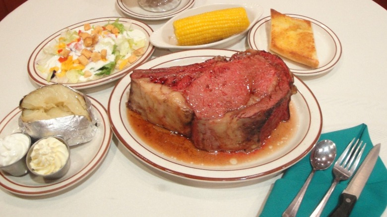 Prime rib on plate with other plates of salad, corn, cornbread, and baked potato