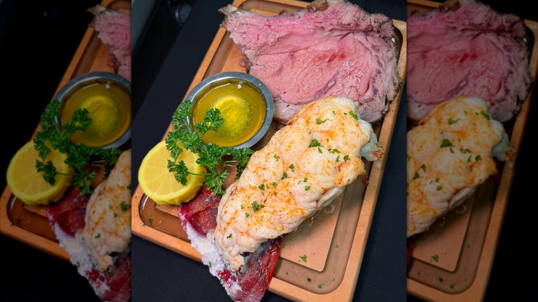 Slice of prime rib and lobster tail on wooden board with lemon and butter