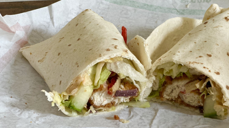 Paper wrapper with wrap cut in half with visible fried chicken, cucumber, tortilla strips, and lettuce