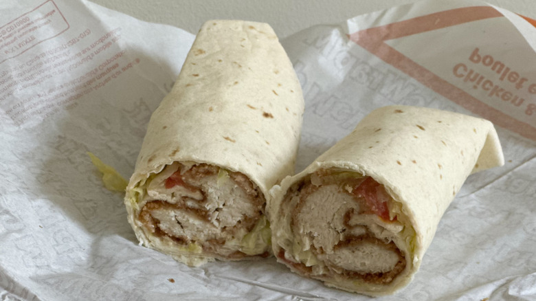 Paper wrapper with wrap cut in half with visible fried chicken, tomato, and lettuce