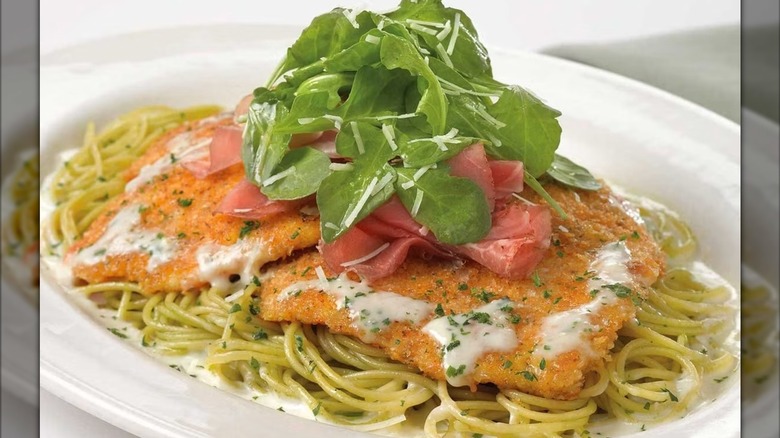 Chicken Bellagio over pesto pasta served on a white plate topped with greens, prosciutto, ad grated cheese at The Cheesecake Factory