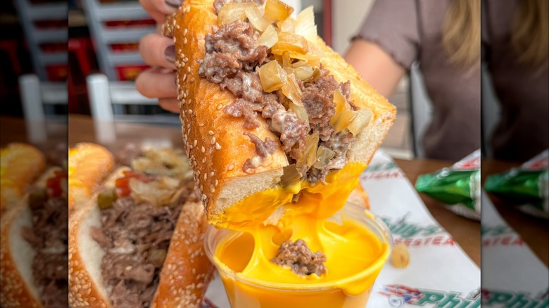 Hand holding cheesesteak dipping into cup of Cheez Whiz