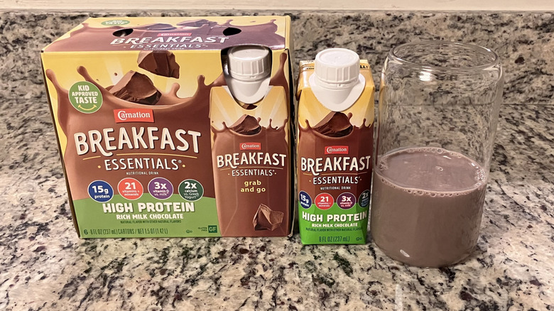 A four-pack of Carnation Breakfast Essentials High Protein Rich Milk Chocolate Shakes sits next to a bottle and clear glass containing the shake