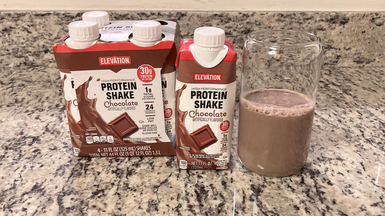 A four-pack of Elevation Chocolate Ready to Drink Protein Shakes sits next to a bottle and clear glass containing the shake